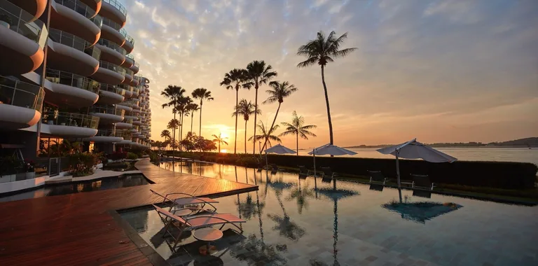 Seascape, photograph: the pool deck at sunset, with sun loungers, parasols and palm trees mirrored in the still water beside the curved balconies