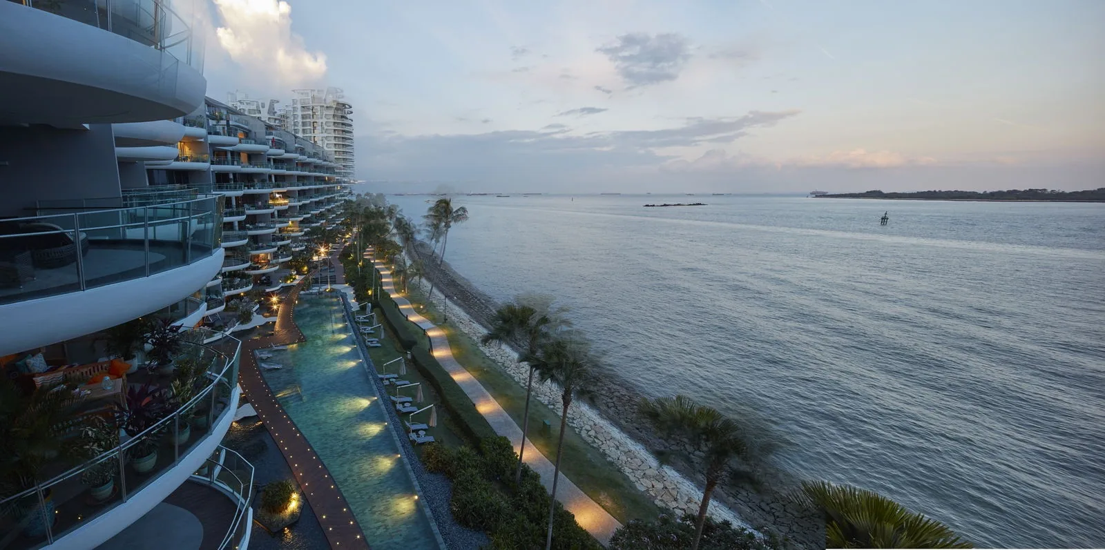 Seascape, photograph: the long pool and its lit terraces running along the shore at dusk, with the curved balconies of the block on the left and the sea and the southern islands on the right